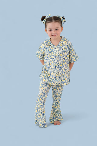 Girl wearing yellow and blue toile print short-sleeve button-up shirt with matching flared pants and coordinating hair bows.