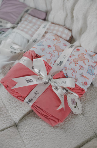 Pink and white ruffled blankets with boot and floral prints, wrapped with white ribbon bands on cream bedding.