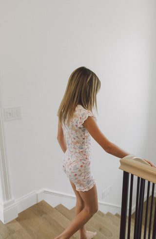 Child wearing white short set with floral and boot print pattern and ruffled sleeves.