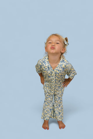 Young child wearing a yellow and blue floral print short-sleeve button-up shirt and matching flare pants against a blue backg