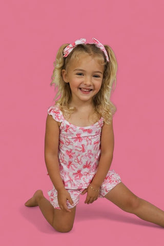 Child wearing pink and white bow-patterned ruffle shorts with matching hair bows against pink background.