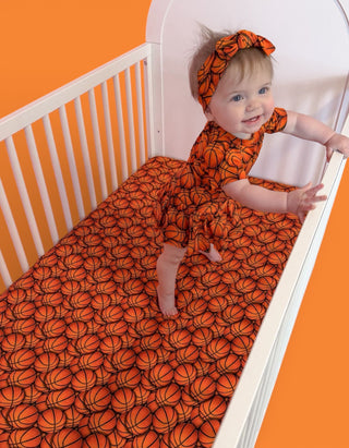 Infant in white crib with orange and black basketball-patterned crib sheet and matching headband.
