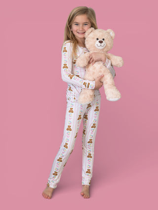 Child wearing white pajamas with bear and floral print, holding matching cream-colored plush teddy bear.