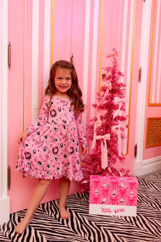Young girl wearing pink patterned dress standing next to pink tinsel Christmas tree and coordinating gift box against striped