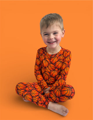 Young child wearing orange long-sleeve pajamas with basketball print pattern, seated against orange background.