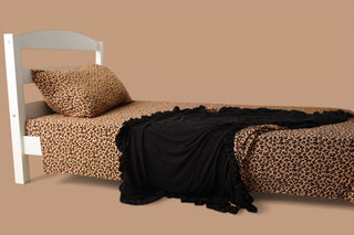 Twin bed with tan leopard print sheets, pillowcase, and matching comforter with black blanket draped across it.