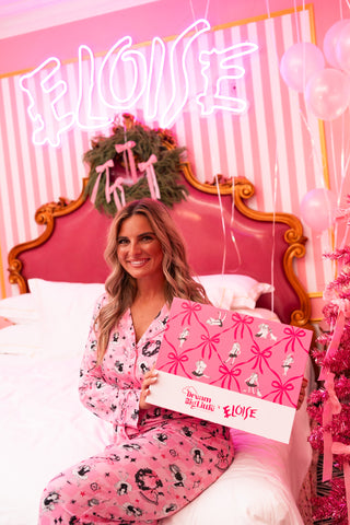 Woman holding pink gift box on bed with neon "ELOISE" sign and decorative pink striped backdrop.