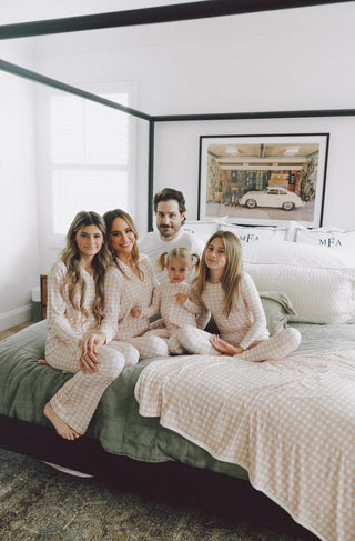 Family wearing matching beige and white gingham check flare pajama sets on bed.