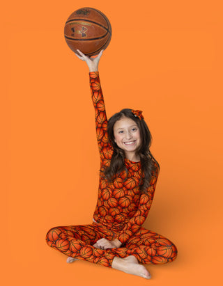 Child wearing orange basketball-print pajamas holding up a brown basketball against orange background.