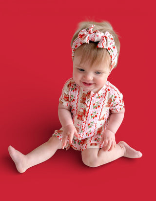 Infant sitting against red background wearing short-sleeved romper with red berry print and matching headband with bow.