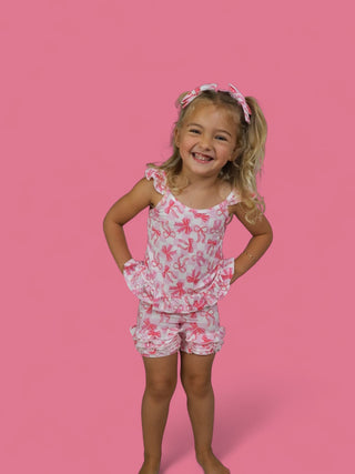 Young girl wearing white shorts with pink bow pattern and matching ruffled tank top against pink background.