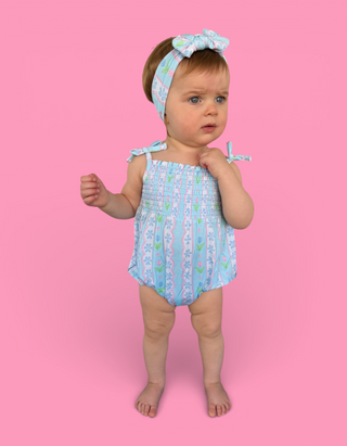 Toddler wearing light blue bubble romper with tulip print and matching headband with bow against pink background.