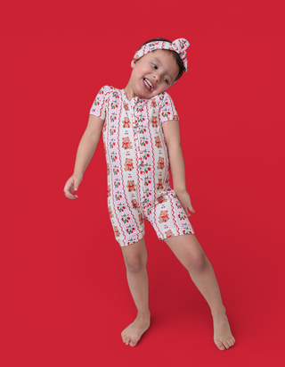 Child wearing white short-sleeved romper with red bear and berry print, matching headband, posed against red background.