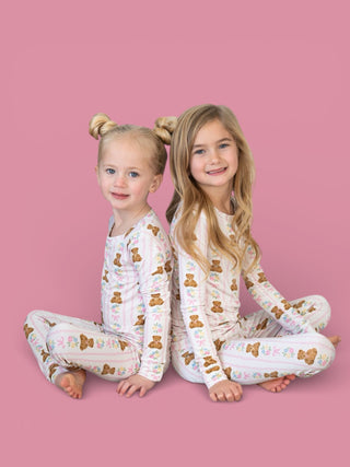 Two young girls wearing matching long-sleeve white pajama sets with brown teddy bears and floral print, seated on pink backgr