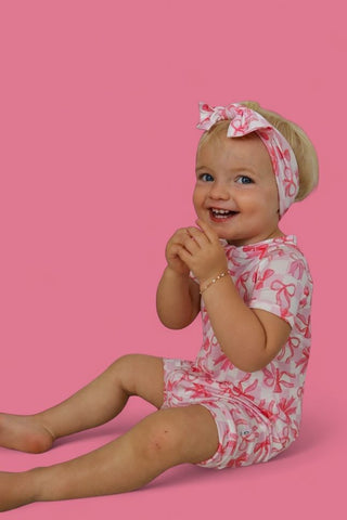 Toddler wearing pink and white checkered bow romper with matching headband against pink background.