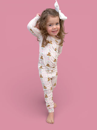 Young child wearing white long-sleeve top and matching leggings with bear and floral print, white bow hair clip, against pink