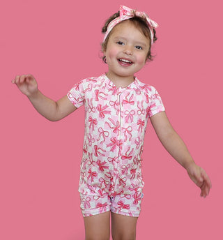 Toddler wearing pink and white bow-print short-sleeve romper with matching headband against pink background.