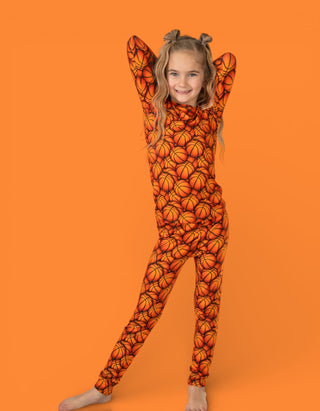 Child wearing orange long-sleeve pajamas with basketball print pattern, posed with arms raised against orange background.