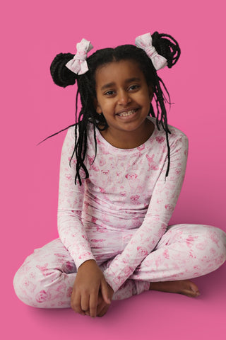 Child wearing pink toile puppy print long-sleeve shirt and matching pants with pink bows in hair against pink background.