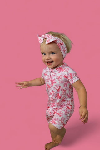 Toddler wearing pink and white bow-print romper with matching headband bow against pink background.