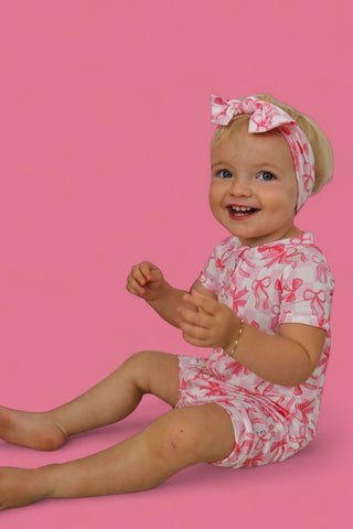 Toddler wearing pink and white bow-print short-sleeve outfit with matching bow headband against pink background.