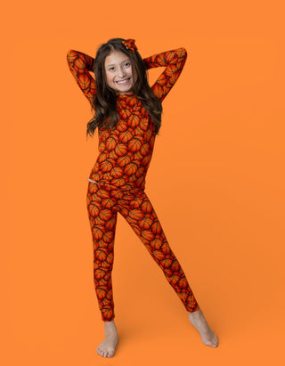 Child wearing orange long-sleeve top and matching leggings with basketball print pattern, hair bow, posed against orange back