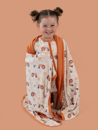 Young child wrapped in cream blanket with orange trim featuring farm animals and tractor patterns.