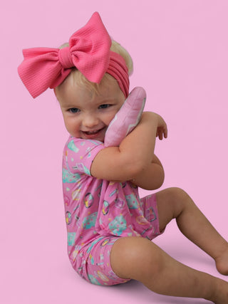 Doll with blonde hair and pink bow headband holding a pink quilted plush toy against pink background.