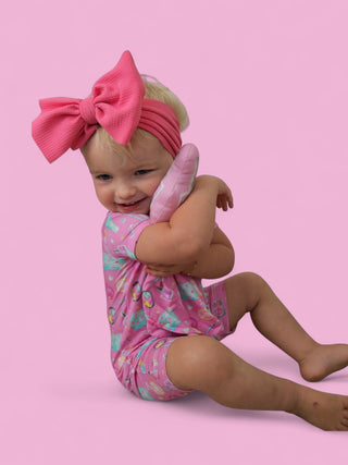 Pink plush lovey with quilted texture held by toddler wearing pink bow headband against pink background.