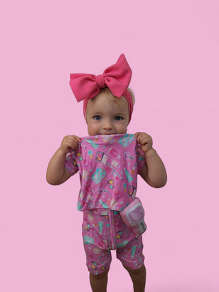 Toddler holding pink plush lovey with colorful pattern, wearing matching outfit and pink bow headband against pink background
