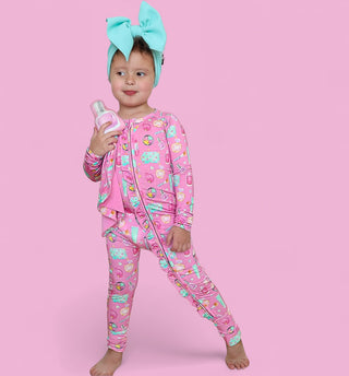 Young child wearing pink pajamas with colorful pattern and turquoise bow headwrap, holding small plush toy against pink backg