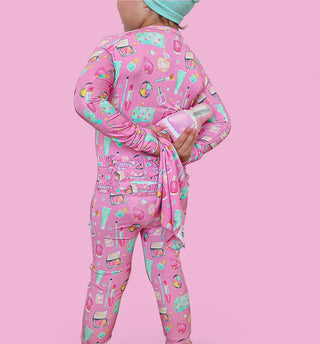 Child wearing pink pajamas with multicolored beauty and cosmetics print, long sleeves, and ruffled waistband detail.