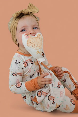 Infant holding a white plush lovey with chicken design next to face, wearing cream pajamas with farm animal and bicycle print