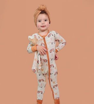 Toddler holding tan plush farm animal toy while wearing cream pajamas with farmyard print and orange trim.