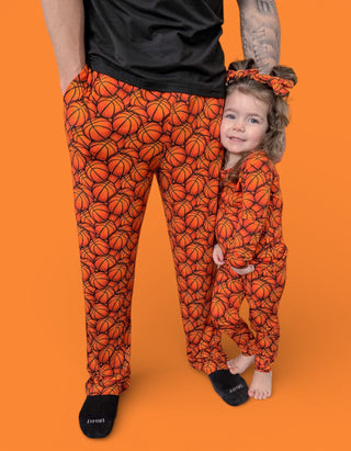 Adult and child wearing matching orange pajama pants with basketball print pattern and black socks against orange background.