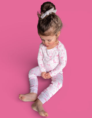 Toddler wearing white pajamas with pink puppy pattern and matching hair bow against pink background.
