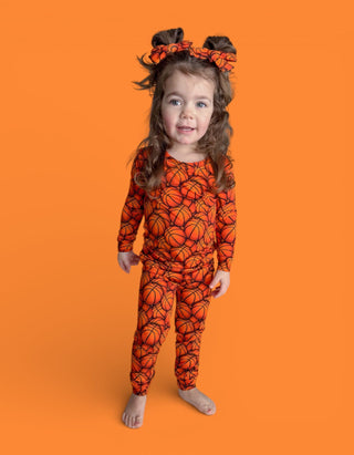 Toddler wearing orange basketball-print long-sleeve shirt and matching pants with coordinating hair bows against orange backg