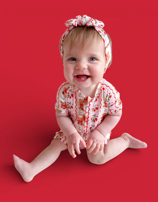 Smiling baby wearing red and white berry-patterned short-sleeved romper with matching headwrap on red background.