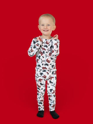 Toddler wearing white pajamas with hockey sticks and skates print, paired with black socks, against red background.