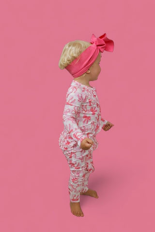 Infant wearing pink checkered bow headband and white romper with pink bow pattern and ruffled legs against pink background.