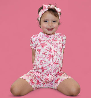 Toddler wearing pink and white bow-print short-sleeve romper with matching headbow against pink background.
