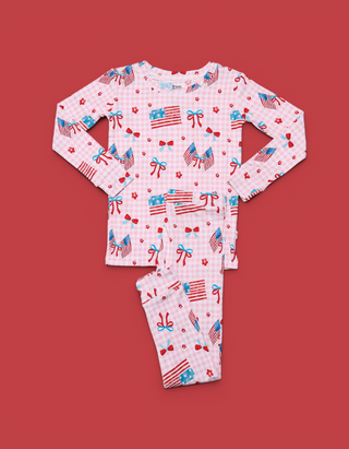 Light pink children's pajama set with long sleeves and pants featuring American flags, red bows, and stars on red background.