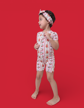 Child wearing short-sleeved romper with berry and bear print, matching headband, standing against red background.