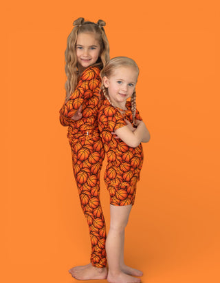 Two children wearing matching orange basketball-patterned pajamas with long sleeves and capri pants on orange background.