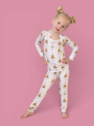 Child wearing white long-sleeve pajama set with teddy bear and floral print pattern against pink background.