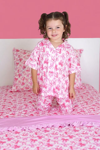 Child wearing pink bow-print pajamas on matching twin sheet set with coordinating pillows.