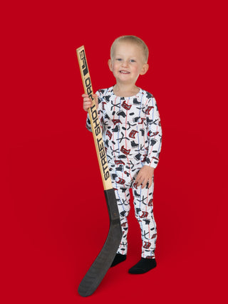 Young child wearing hockey and skateboard-patterned pajamas holding a wooden hockey stick against red background.