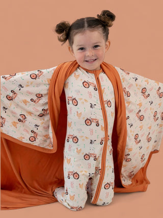 Cream blanket with farmyard animal and tractor print, orange trim and backing, displayed on toddler.