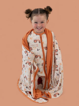 Toddler wrapped in cream blanket with orange trim, featuring farm animal and tractor print design.