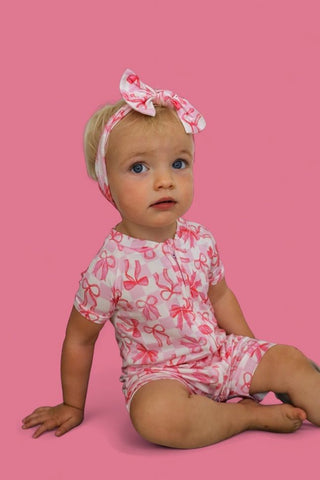 Infant wearing pink and white bow-patterned short-sleeve romper with matching bow headband against pink background.